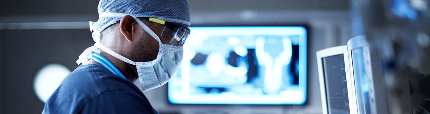 Male surgeon wearing surgical mask and safety glasses looks a monitor, image from blurred monitor fills background