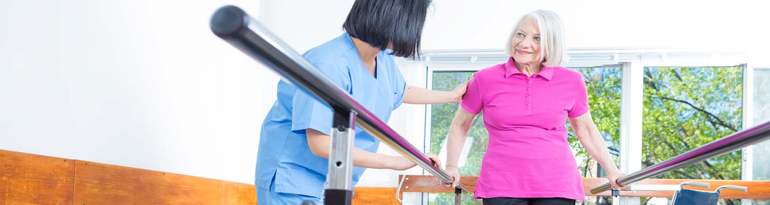 Senior woman using parallel bars to walk, smiling looking at nurse who is aiding her